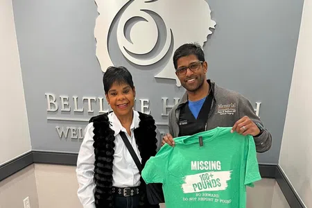 Two people standing in Beltline Health office, one holding a green shirt saying 'Missing 100+ Pounds' smiling