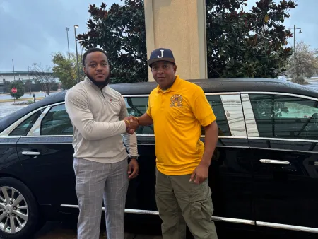 Two men shaking hands and posing beside a black car in an outdoor setting with trees and pavement.