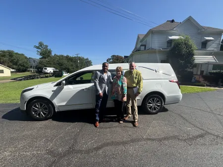 Three people standing in front of a white luxury hearse vehicle on a sunny day near a funeral home.