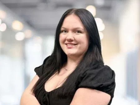 Smiling woman with long black hair wearing a black dress standing confidently with arms crossed in bright office.