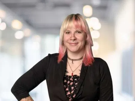 Woman with pink ombre hair, tattoos, and black blazer smiling confidently in a blurred office background