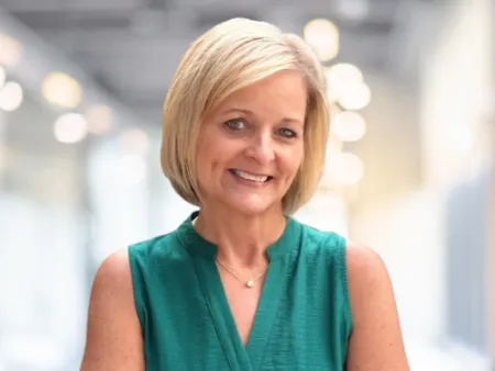 Smiling middle-aged woman with short blonde hair wearing a sleeveless green blouse in a bright office setting