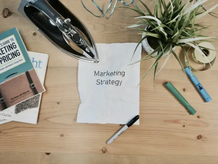 Workspace with marketing books, iron, plant, and a crumpled paper labeled Marketing Strategy on wooden table.