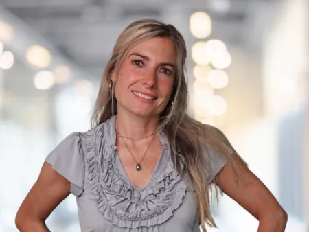 Smiling woman with long blonde hair wearing a gray ruffled blouse in a bright indoor setting.