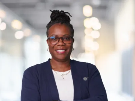 Smiling woman with glasses and braided hair wearing a navy cardigan and smartwatch standing with arms crossed indoors
