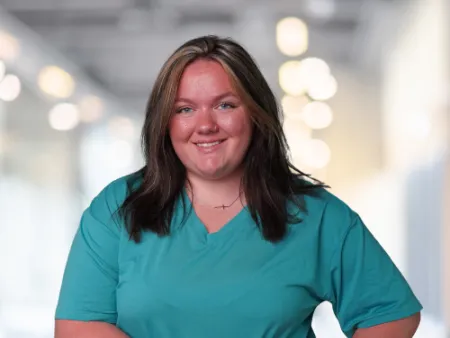 Smiling woman in teal scrub top standing in bright medical or office hallway with blurred lights behind her