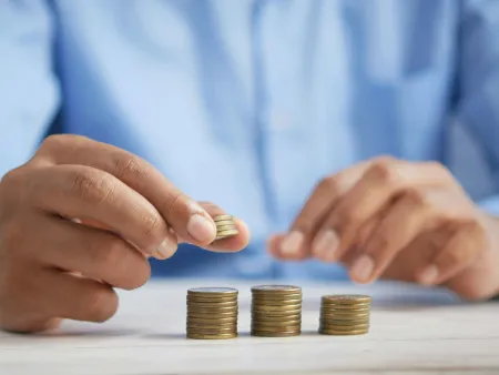 Person stacking coins into three small piles on a white surface with a blurred blue shirt background.