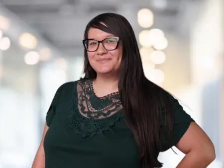 Confident woman with long dark hair and glasses wearing a dark green top with lace detail in a bright office.