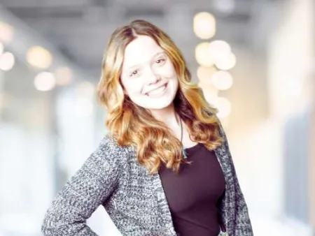 Young woman with long wavy hair in gray cardigan and jeans smiling indoors with blurred lights background