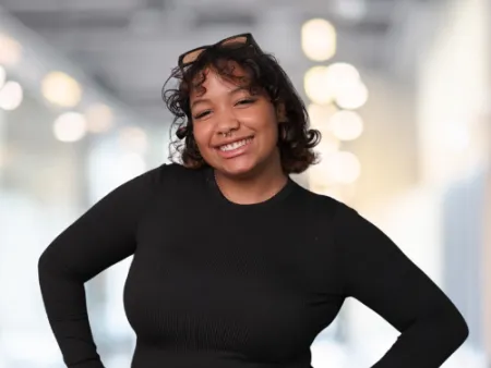 Smiling young woman with curly hair wearing a black long-sleeve shirt and blue jeans in a bright indoor setting