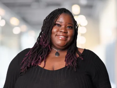 Smiling woman with purple-tipped dreadlocks wearing a black top and pendant necklace in a bright indoor setting