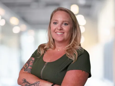 Smiling woman with blonde hair and tattoos wearing a green dress standing with arms crossed in bright modern office.