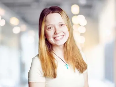 Smiling young woman with long blonde hair wearing a white t-shirt and blue cross necklace indoors