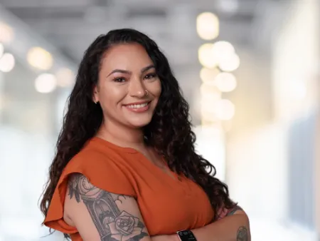 Smiling woman with curly hair, tattoos on arms, wearing orange blouse and smartwatch, standing with arms crossed indoors.