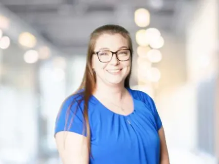 Smiling woman in blue shirt and glasses standing in bright modern office interior with blurred background