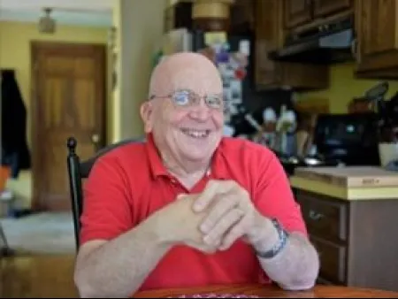 Smiling elderly man in a red polo shirt sitting at a kitchen table with hands clasped relaxed