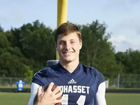 Smiling football player in a blue Cohasset jersey holding a football on a field with goalpost and trees in the background