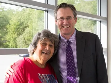 Smiling elderly woman in red shirt with American flag design hugs man in suit near window with green trees outside.