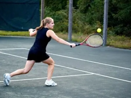 Female tennis player in black dress lunging to hit ball on outdoor clay court surrounded by green trees.