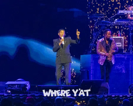 Singer performing on stage with saxophonist and DJ, vibrant lighting and audience visible in foreground