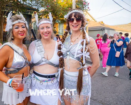 Three women in futuristic silver festival costumes posing outdoors at a sunny street event with a festive crowd.