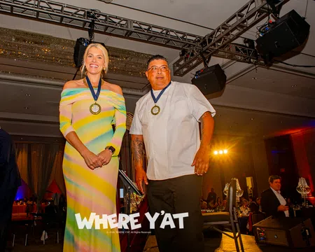 Two award recipients wearing medals stand on stage at a formal event with audience in background.
