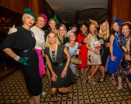 Group of women dressed in vintage-style dresses and hats posing indoors for a lively social event photo.