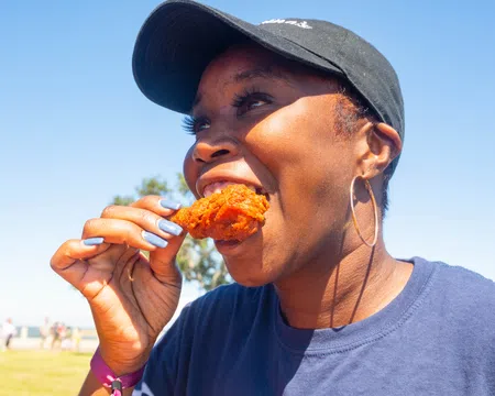 Smiling woman wearing a black cap and hoop earrings eating fried chicken outdoors on a sunny day