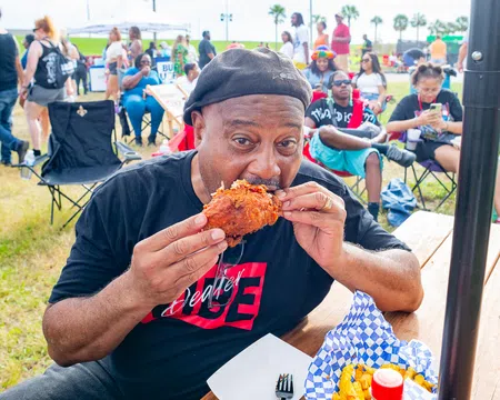 Man in black hat eating fried chicken at outdoor event with people and tents in the background