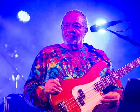 Musician wearing a colorful tie-dye shirt playing red electric bass guitar on stage with blue lighting.