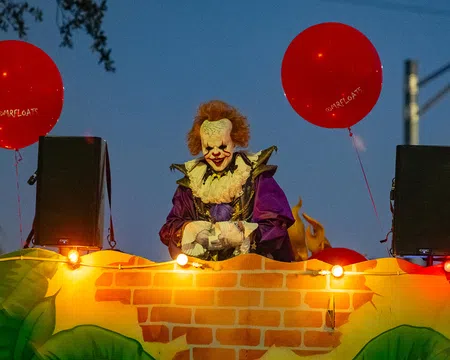 Costumed clown with red hair and white face paint holding red balloons at night during parade float event.