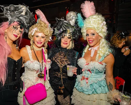 Four women in elaborate Halloween costumes with wigs and accessories at a festive party indoors.