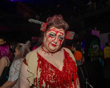 Woman in bloody costume with fake cleaver through head at a Halloween party with crowd in background