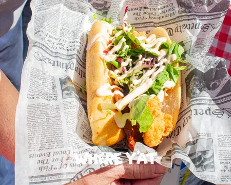 Fresh fried fish sandwich with lettuce and sauce served in newspaper-style wrapping held by a person.