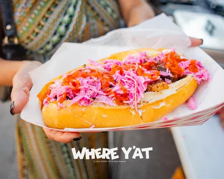 Close-up of a hand holding a hot dog with pink slaw and red sauce in a paper tray, with overlaid text reading WHERE Y'AT.