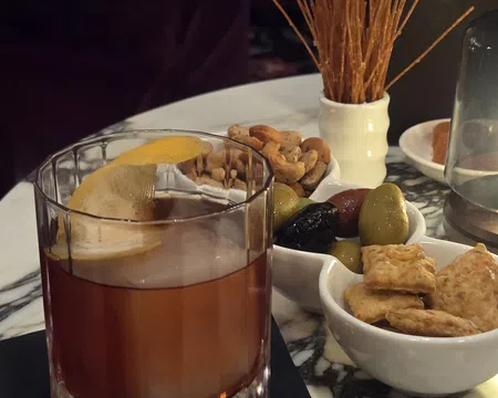 Old fashioned cocktail with lemon peel served with assorted nuts, olives, crackers, and breadsticks on marble table.