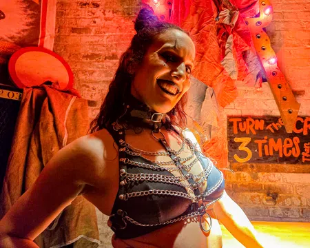 Woman in black leather harness and chains smiles against a brick wall with Halloween decor and overlaid text sign.