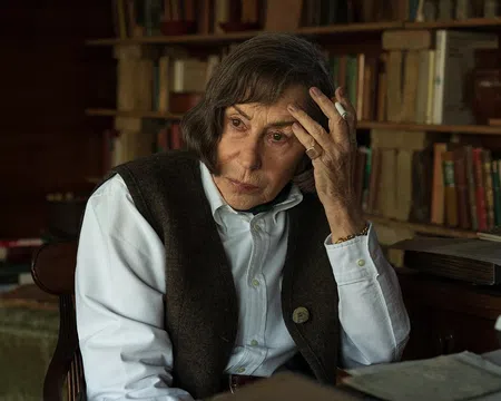 Elderly woman with brown hair sitting contemplatively in a vintage study room filled with books and a typewriter.