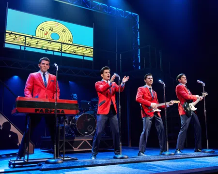 Four male performers in red jackets play instruments and sing on stage with musical notes backdrop in blue lighting.