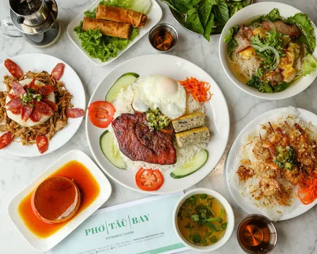 Overhead view of Vietnamese dishes including grilled pork with egg, spring rolls, noodle soup, shrimp skewers, and caramel flan on a marble table.