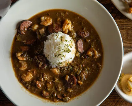 Bowl of shrimp and sausage gumbo with white rice and herbs on top, served on a wooden table with side dishes.