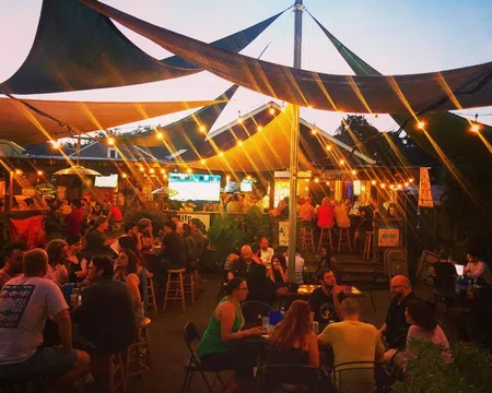 Outdoor evening bar scene with string lights, shaded canopies, and groups of people socializing under warm lighting.