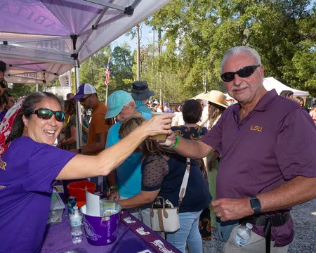 People enjoying an outdoor event exchanging drinks under purple tents on a sunny day with trees in the background