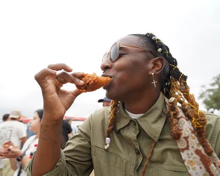 Person with braided hair and sunglasses enjoying crispy fried chicken outdoors at a casual event.