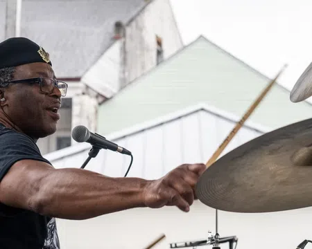 Man wearing a beret playing drums energetically at an outdoor performance with houses in the background