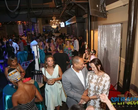 Crowded lively indoor party with guests socializing, dressed in formal and cocktail attire under chandeliers.