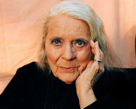 Elderly woman with silver hair, wearing black, resting her face on her hand against a soft peach background.
