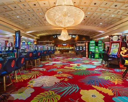 Colorful casino interior with floral carpet, gaming machines, blue chairs, and large chandeliers overhead