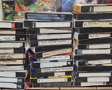 Stack of vintage VHS tapes with handwritten labels on a cluttered desk surrounded by posters and household items.