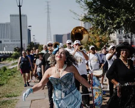 Woman in blue corset and gloves dances joyfully in front of a marching band and parade crowd on sunny day.